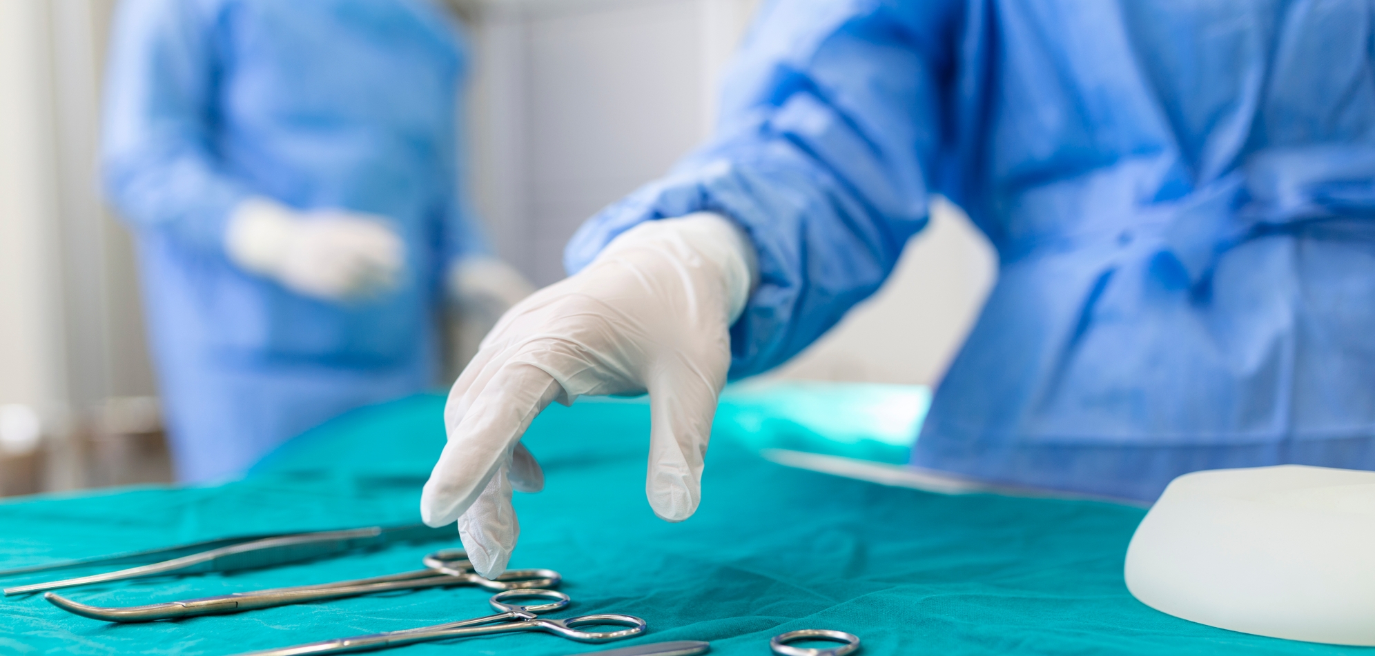 Nurse hand taking surgical instrument for group of surgeons at background operating patient in surgical theatre. Steel medical instruments ready to be used. Surgery and emergency concept Nurse hand taking surgical instrument for group of surgeons at background operating patient in surgical theatre. Steel medical instruments ready to be used. Surgery and emergency concept